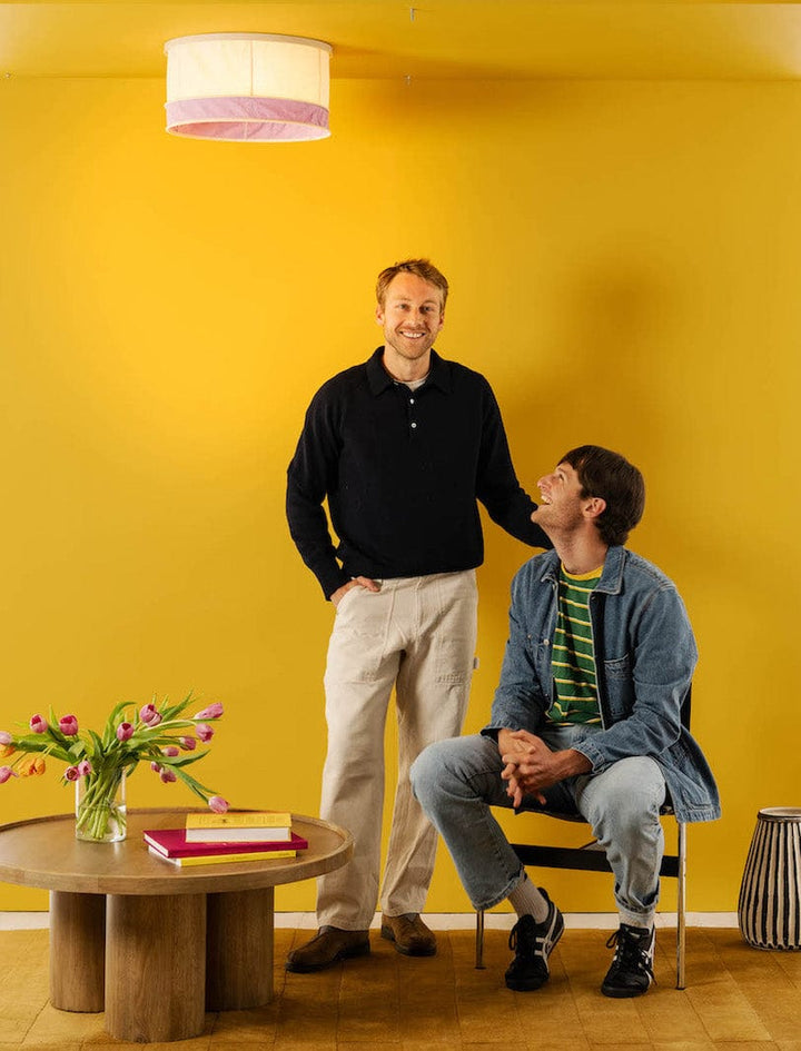 Two men standing under a lilac, drum shaped ceiling light cover. They are standing next to a table with a vase of tulips on top of it.